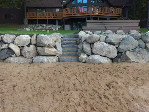 Boulder retaining wall with stone steps leading down to a sandy beach below a deck