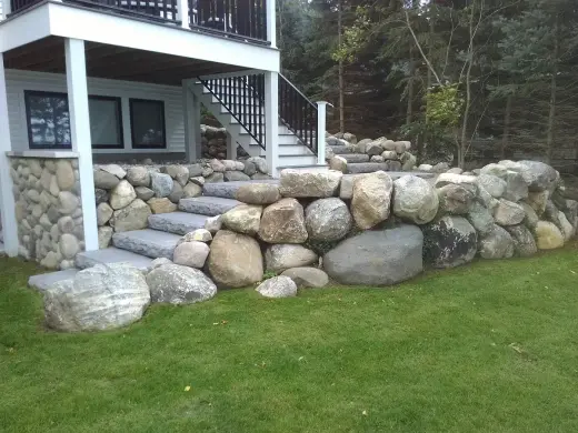 Large boulder retaining wall with stone slab steps below an elevated white deck