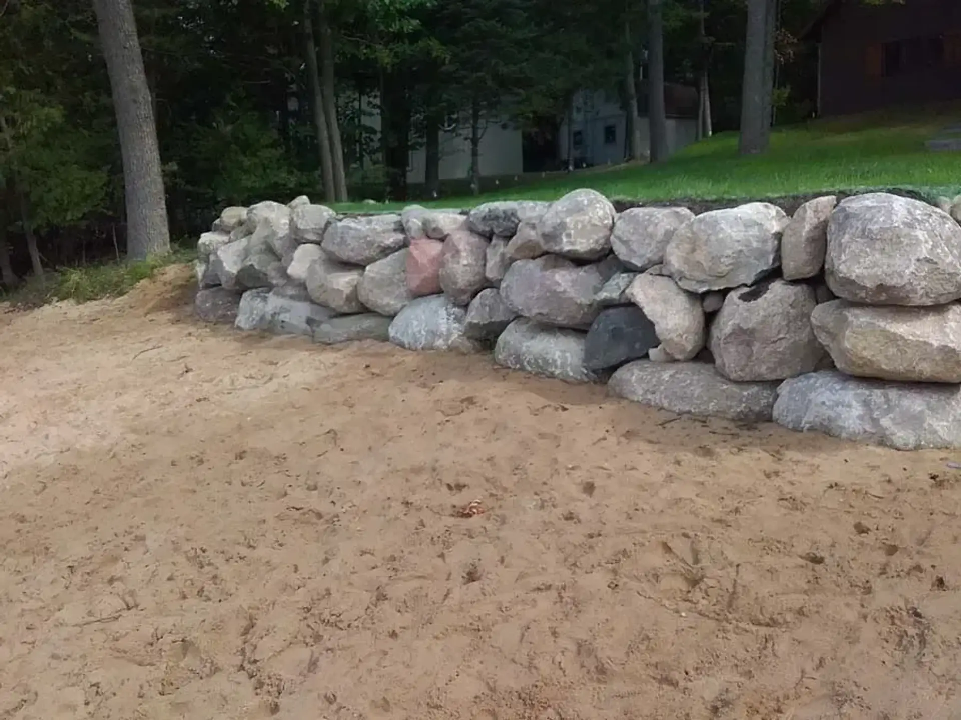 Large fieldstone boulder retaining wall along leveled sandy ground in a wooded yard