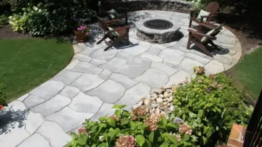 Round fire pit on a curved stone patio with wooden chairs, hydrangeas and lawn