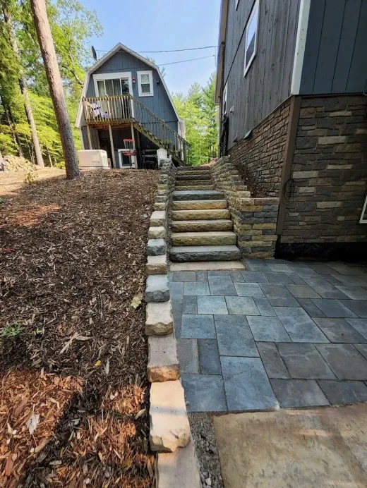 Gray paver patio with natural stone steps and a stone wall beside a dark-sided home