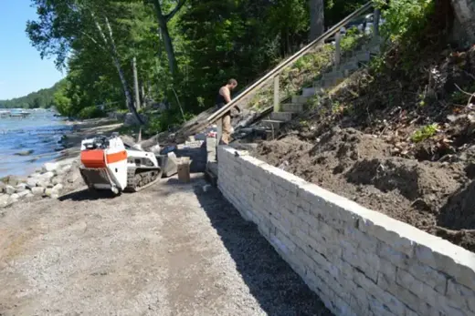Tan stacked block retaining wall at a lakeshore with a skid steer on gravel and wood stairs