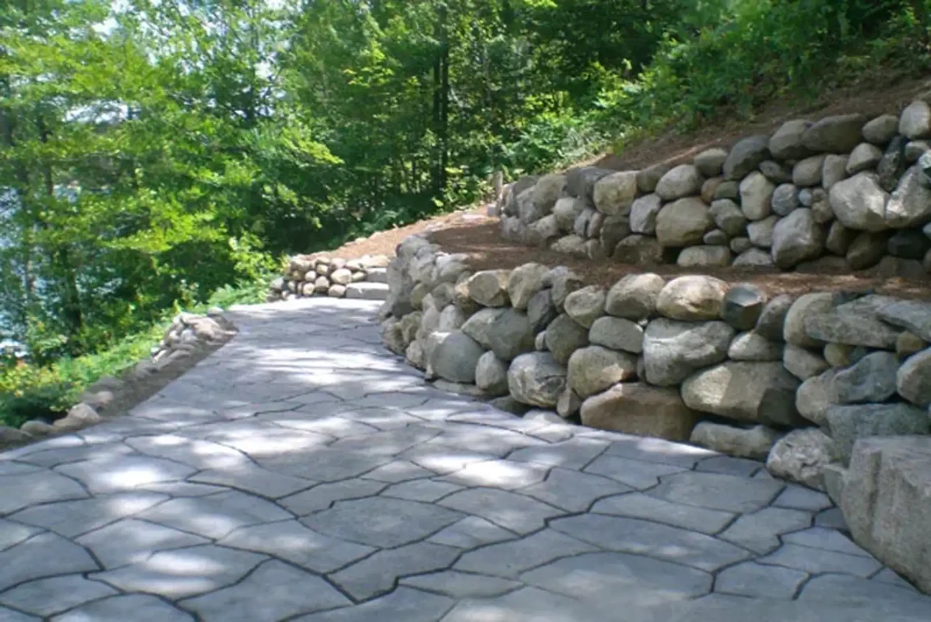 Stamped concrete walkway curving beside two-tier boulder retaining walls on a wooded slope