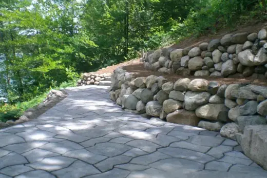 Stamped concrete walkway curving beside two-tier boulder retaining walls on a wooded slope