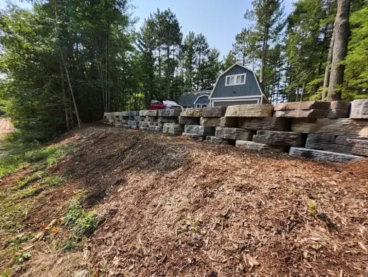 Stacked natural stone retaining wall above a mulched slope near a gray gambrel garage