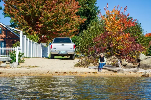 Person sitting on a sandy lakefront beach with fall foliage and a pickup truck behind