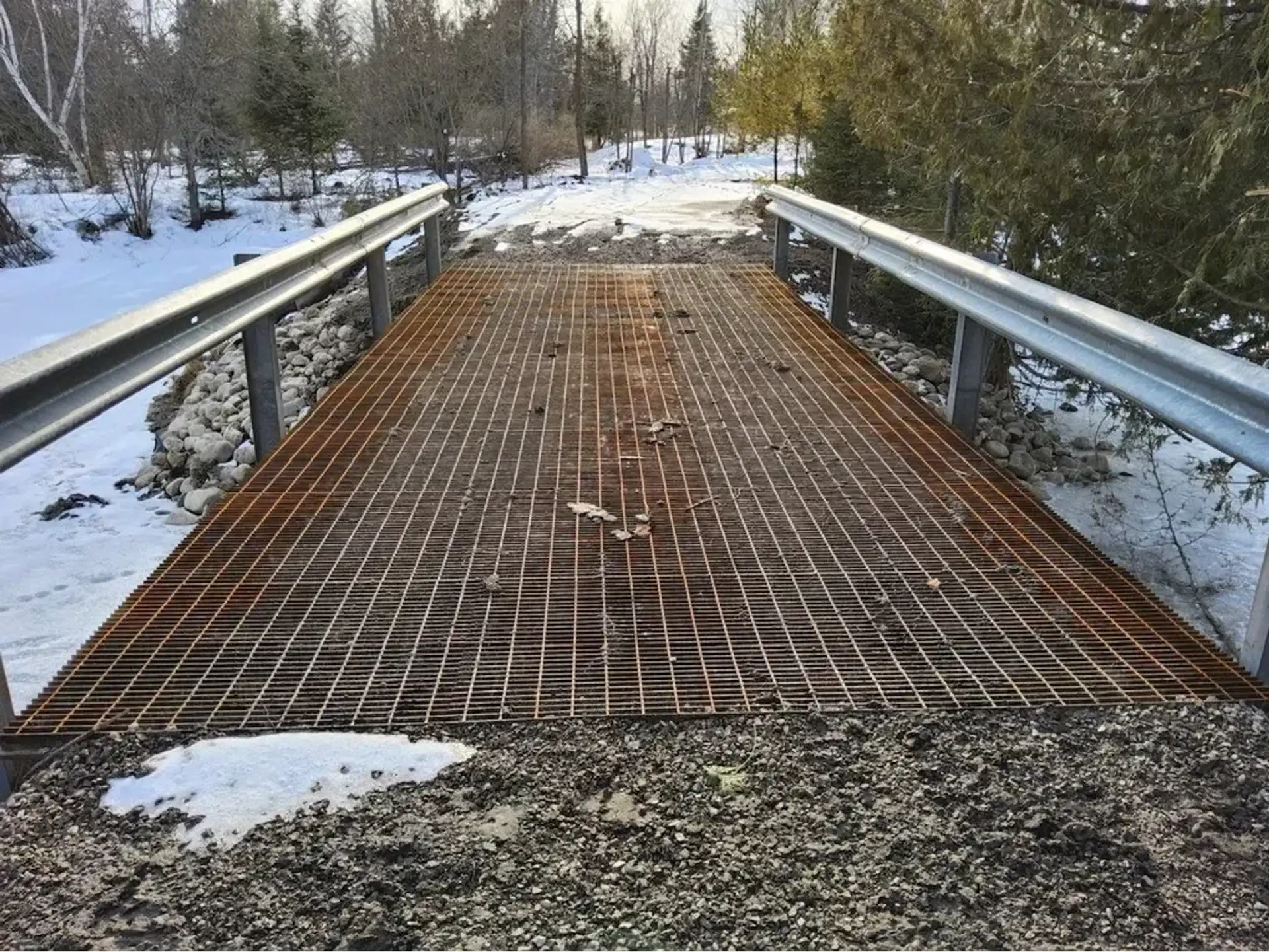 Steel grate bridge deck with guardrails over a gravel approach in snowy woods
